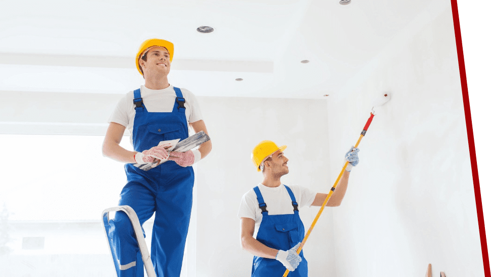 Two men in blue overalls and hard hats are holding paint brushes.