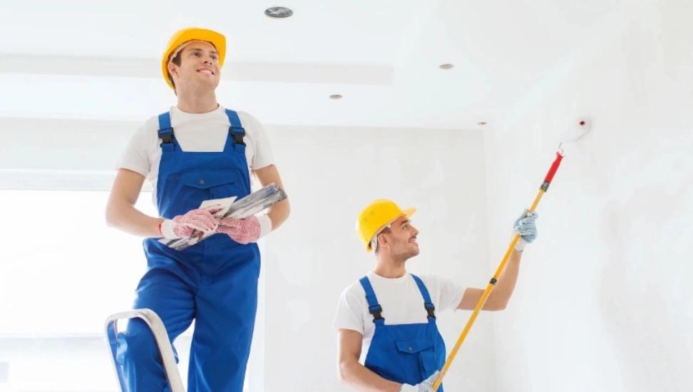 Two men in blue overalls and hard hats are painting a room.